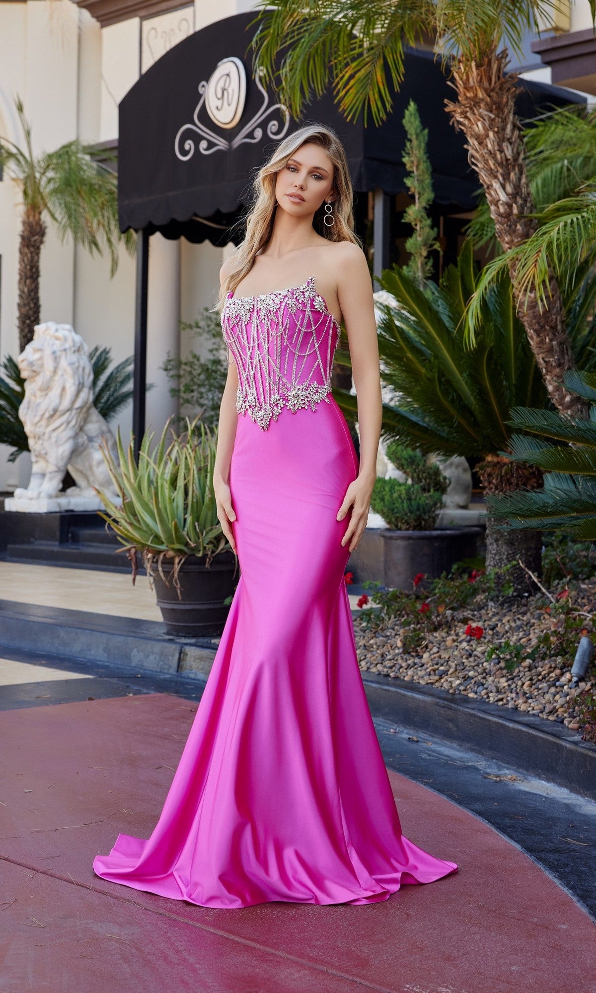 A woman wears the Nox Anabel E1901 Long Prom Dress in fuchsia, featuring a strapless, fitted bodice and satin skirt with crystal beaded embellishments, standing outdoors near tropical plants and a black awning.